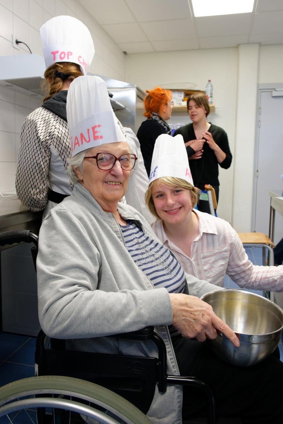 Une personne âgée et une adolescente, toques sur la tête, cuisinent ensemble et sourient lors d'un atelier intergénérationnel de l'association.