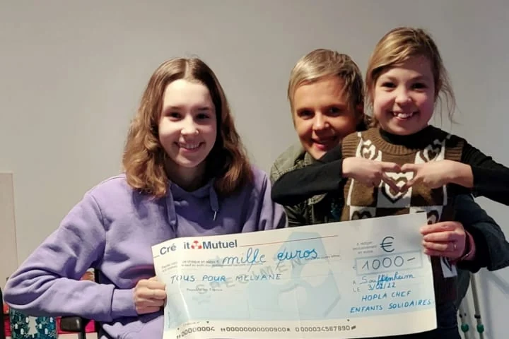 Mélyane, en fauteuil roulant, reçoit un chèque de 1 000 € de l’association, entourée de deux enfants souriants.