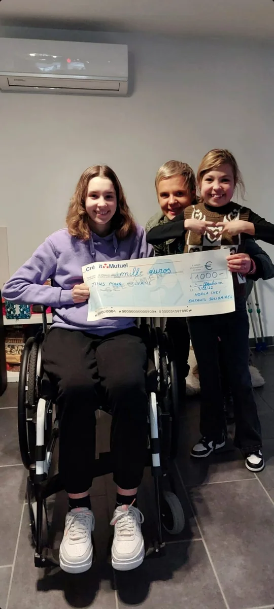Mélyane, en fauteuil roulant, reçoit un chèque de 1 000 € de l’association, entourée de deux enfants souriants.